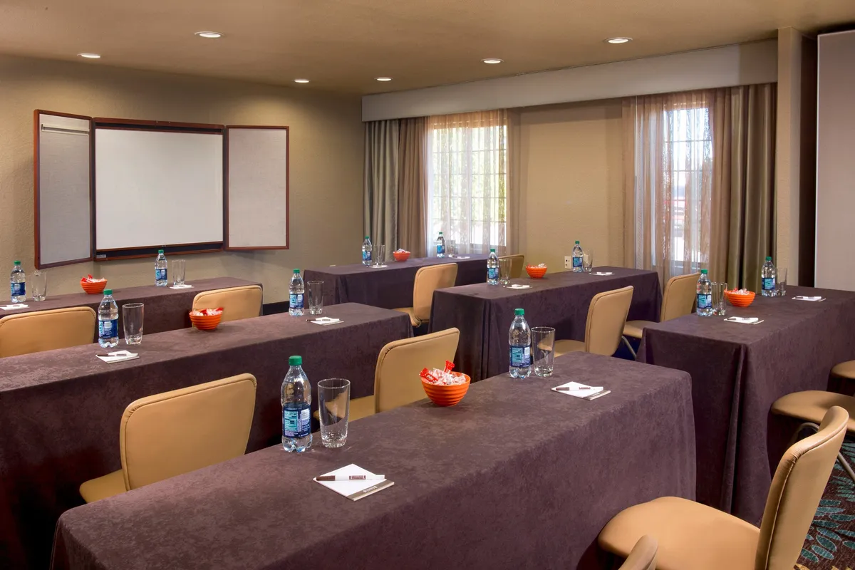 A bright, naturally lit business center setup with tables, water bottles, and a presentation board is showcased. The space is ideal for meetings, workshops, and productive solo work, reflecting a professional, focused atmosphere.