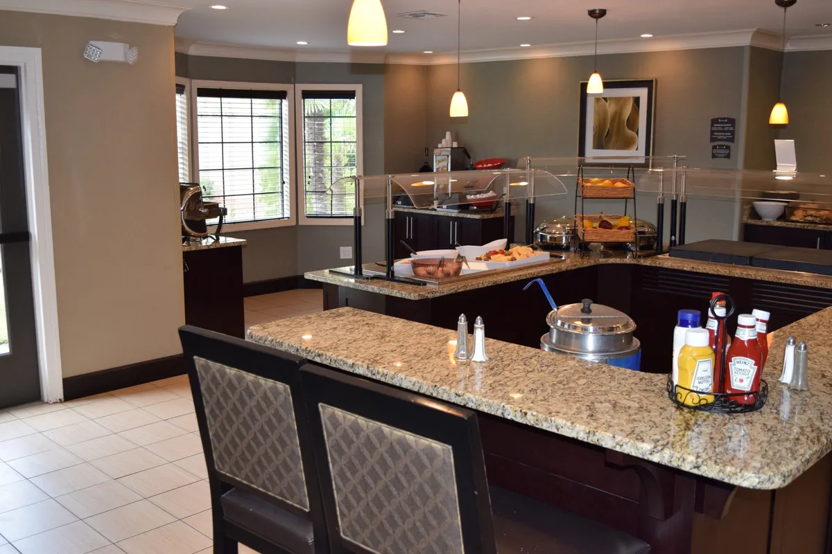 An interior view showing a granite countertop laden with a variety of food items and a condiment station, warmly lit by stylish fixtures. The setup suggests both functionality for meal preparation and a hospitable space for sharing food with loved ones.