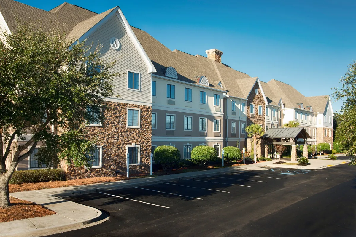 The image shows the main entrance to the property, with a striking canopy, a blend of stone and siding exterior, and lush, landscaped shrubs and trees in the foreground. The welcoming, inviting atmosphere suggests easy access to both the accommodations and the surrounding attractions.