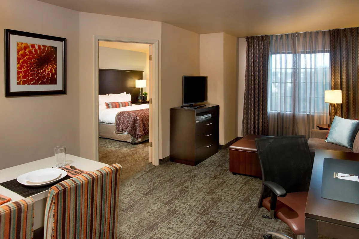 The image showcases a bright, welcoming condo interior with a set dining table ready for a meal, a comfortable bed in the background, a modern television unit for entertainment, and a large window letting in plenty of natural light. The arrangement invites gatherings, relaxation, and a cozy stay, reflecting both comfort and functionality.