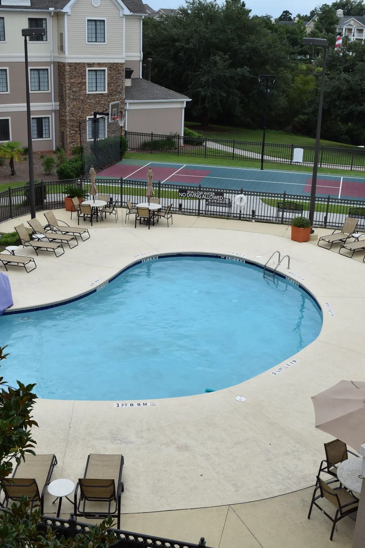 A vibrant exterior scene featuring a swimming pool surrounded by lounge chairs and a nearby tennis court. The area exudes a leisurely, welcoming atmosphere, perfect for sunbathing, swimming, or enjoying evening swims as the sun sets. The pool is central, highlighting the property's outdoor amenities and communal spaces.