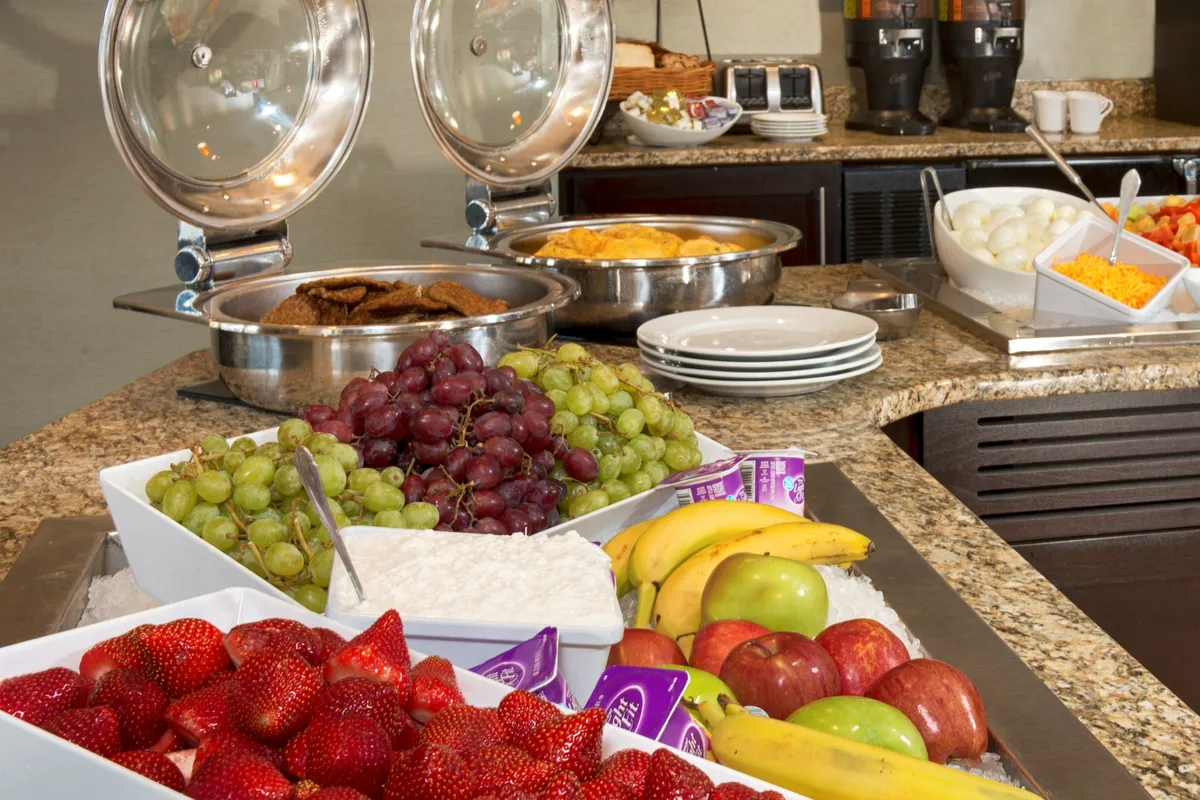 This image captures a vibrant breakfast buffet scene featuring colorful fruit displays, hot food trays, and a well-stocked coffee station. Guests are seen enjoying breakfast and socializing, with an overall cheerful and inviting mood.