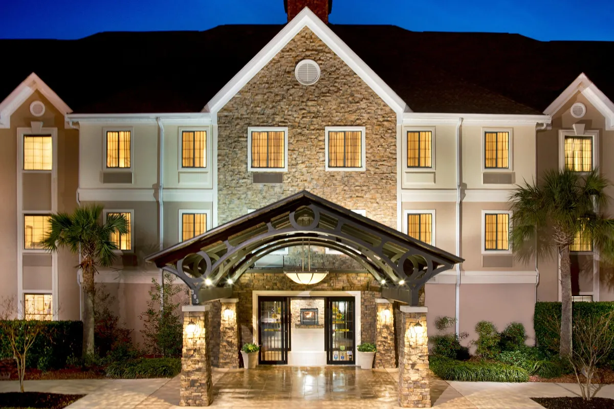 A beautifully landscaped entrance with a stone façade, arched entryway, soft lighting from the windows, and elegant gardens. The scene feels warm and welcoming, inviting guests to begin their getaway in style and comfort.