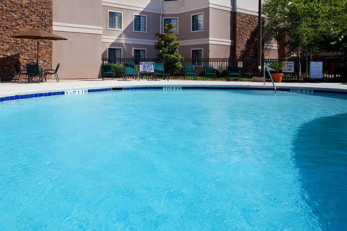 A sun-filled outdoor pool area with a sparkling swimming pool, numerous lounge chairs for sunbathing or relaxing, manicured landscaping, and the welcoming facade of the condo hotel as a backdrop. Couples and guests can be seen enjoying leisurely activities around the pool, suggesting a relaxing, vacation-like atmosphere.