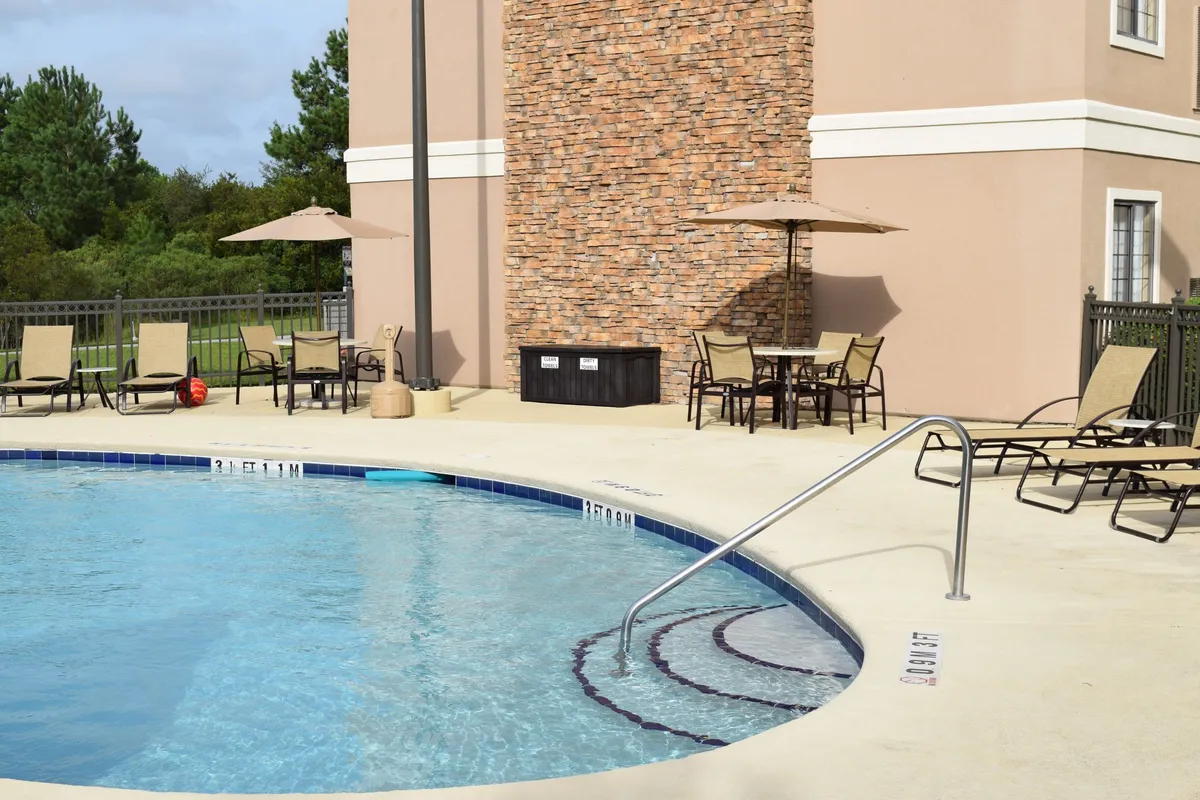 This inviting pool area features a sparkling swimming pool surrounded by lounge chairs and patio umbrellas, set against a warm brick wall. The scene is welcoming, with space for families to relax, swim, and unwind after an active day.