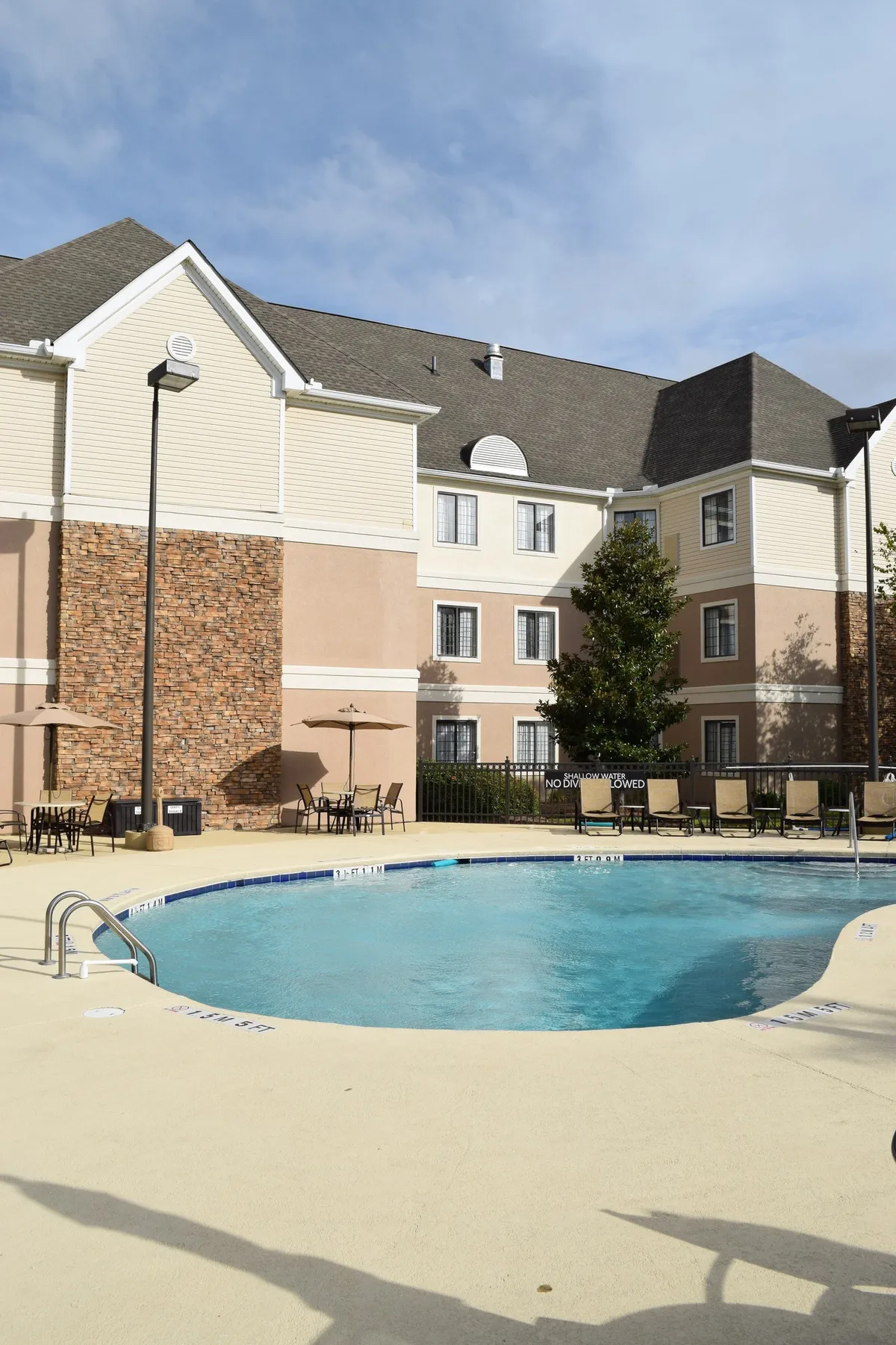 This image shows the building’s exterior with a view of the swimming pool, outdoor seating area, and the facade. There are lounge areas for relaxing and space for socializing, all set in a welcoming, well-maintained environment.