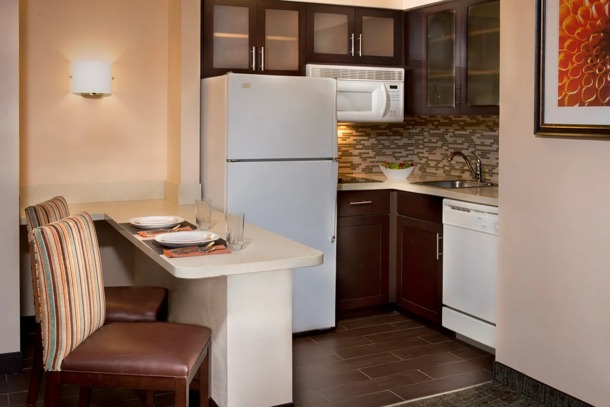 This image features a cozy, well-appointed kitchen with a prominent countertop, a full-size refrigerator, dishwasher, and inviting dining chairs. The scene suggests casual dining and the potential for intimate gatherings or family meals, with subtle hints of activity like cooking and entertaining.