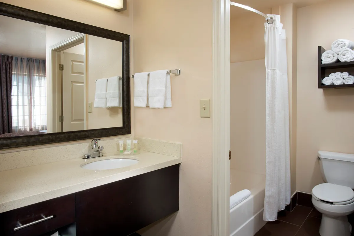 A bright and inviting bathroom interior with a wide sink area, a large mirror, neatly folded towels, and a partially visible shower curtain. The surfaces are clean and minimalist, evoking a sense of calm and order. Soft lighting highlights the tidy setup, suggesting both comfort and privacy.